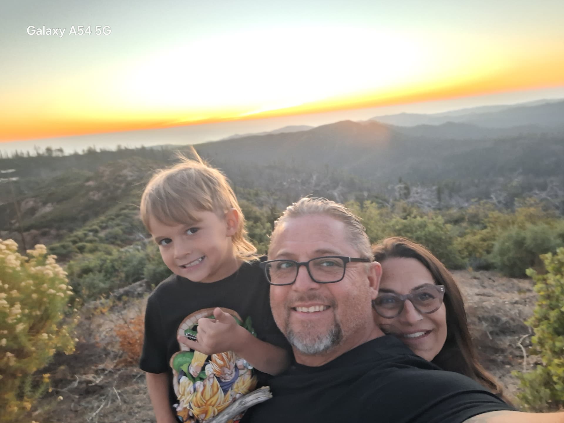 Family selfie of a man, a woman and a young boy smiling outdoors at sunset