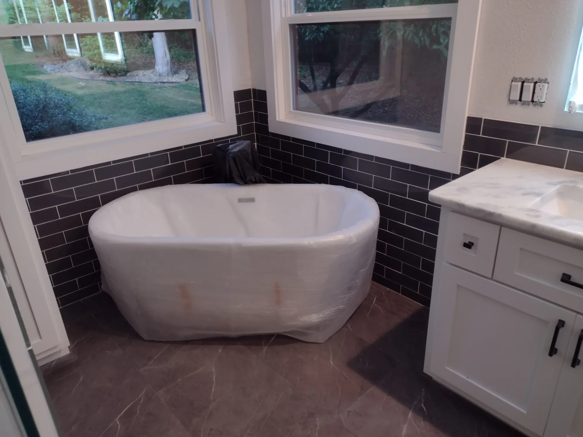 Freestanding tub still in protective wrap, dark subway tile wainscot and white vanity